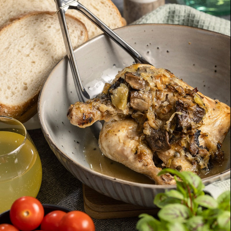 Two Chicken with White Wine, Mushroom & Sage marylands sit in a serving bowl on a table. There are a pair of tongs in the bowl, ready to serve.  Also on the table is some crusty bread, a bowl of cherry tomatoes, a drink and a salt shaker.