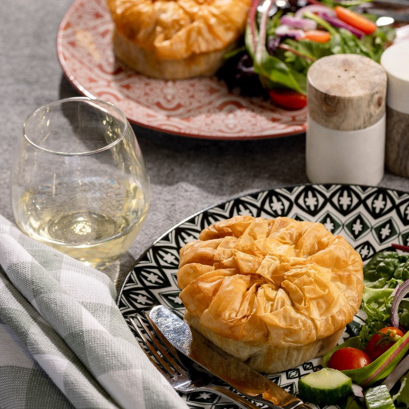 An individual Beef & Vegetable Filo Pie sits on a patterned black and white plate, ready to eat with a knife and fork and side salad.  There is a second pie on a patterned red plate in the background.