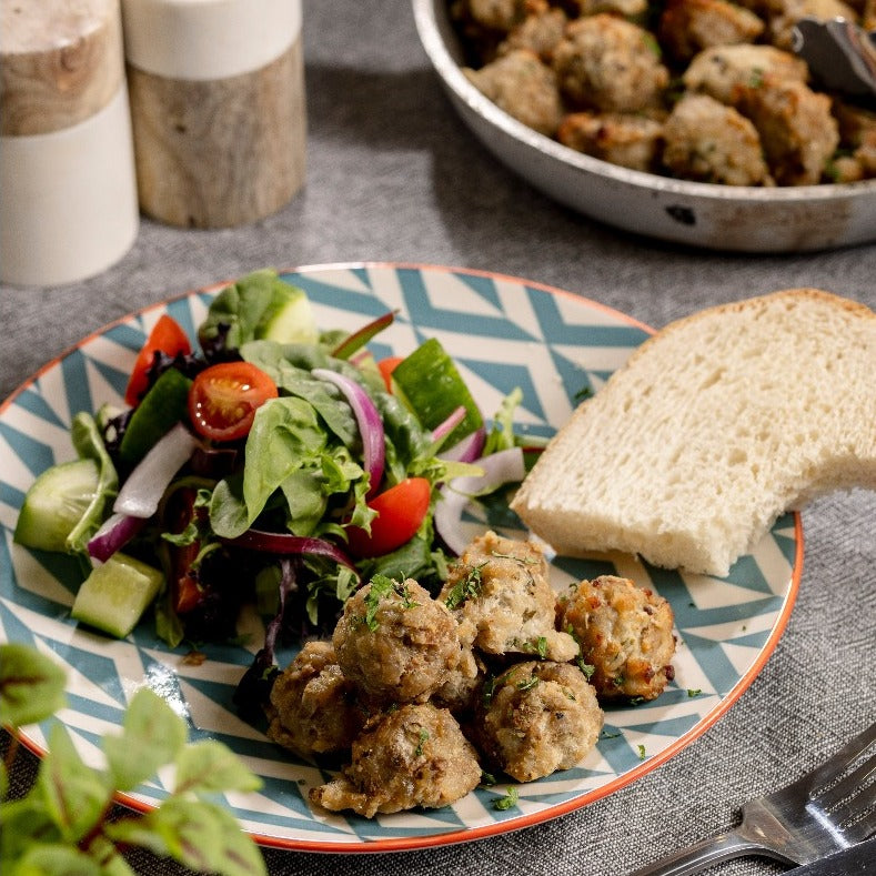 Pork, apple & caramelised onion meatballs sit on a plate with some salad and sourdough bread.  There is a pan of meatballs & salt & pepper shakers in the background.