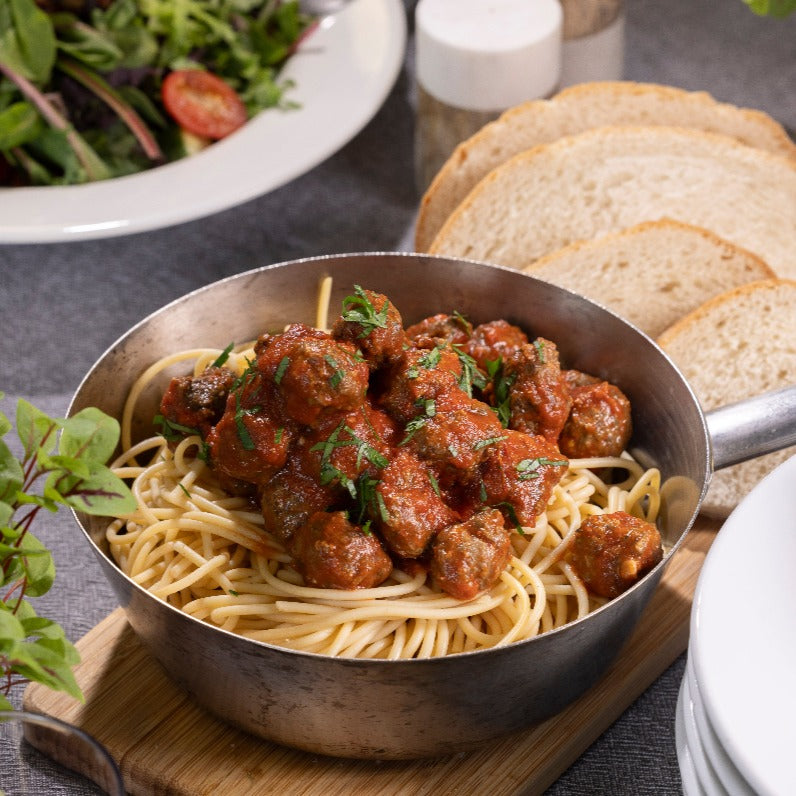 A saucepan of spaghetti with meatballs in classic tomato sauce sits on a wooden board on a table. There is sourdough bread and salad in the background.