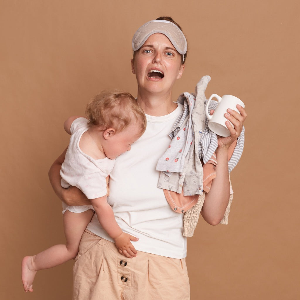 Harassed woman holding a baby, washing on her shoulder and a coffee mug. 