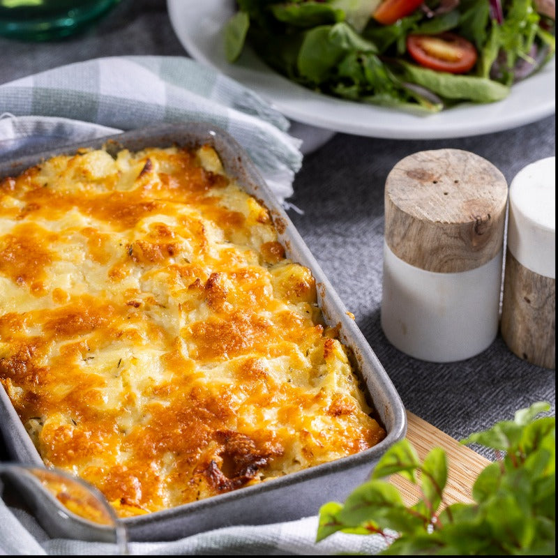 A hot dish of Cauliflower Mac & Cheese sits on a table, a green & white checked cloth wrapped around it.  There is a side salad and salt and pepper shakers in the background.