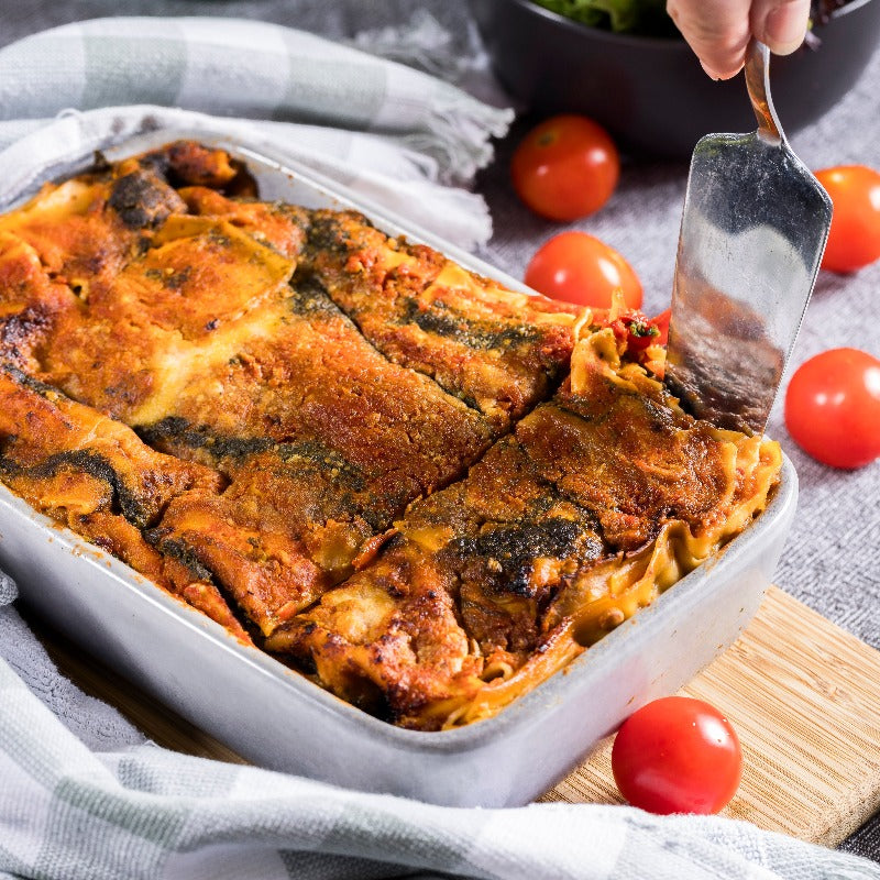 A dish of vegetarian lasagne sits on a chopping board. Someone is using a silver server to scoop out a slice