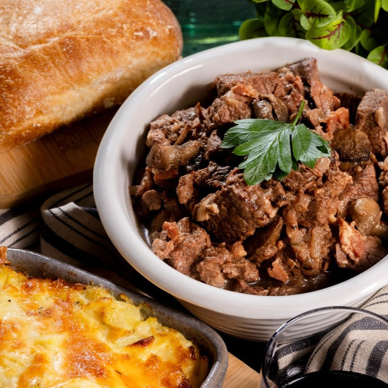 A bowl of Beef Bourguignon sits on a table with a Potato Bake & crusty bread alongside.