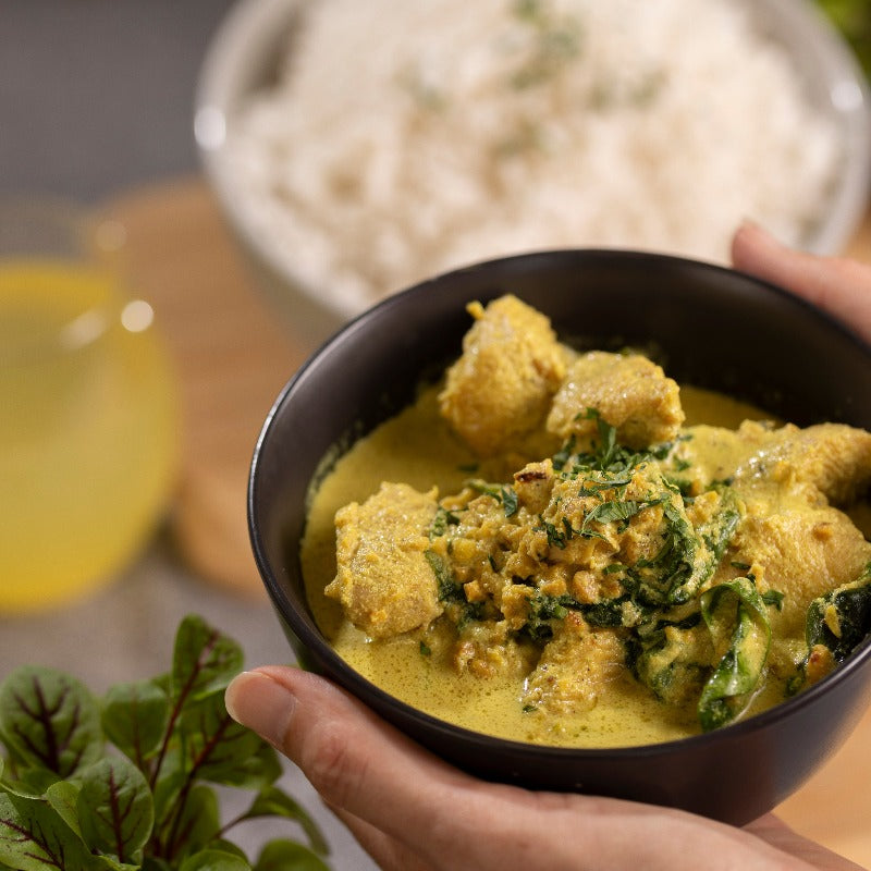 Two hands bring a bowl of Chicken & Peanut Curry to the table.  Already on the table is a bowl of steamed rice and a drink.