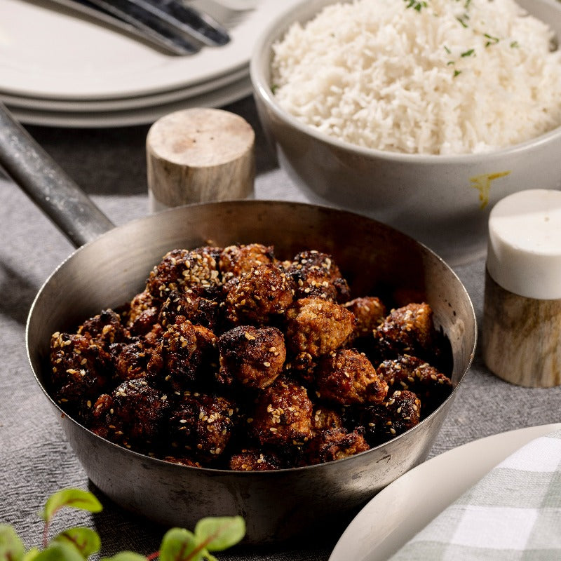 A pan of sesame chicken balls sits on a table. There is a bowl of steamed rice in the background.