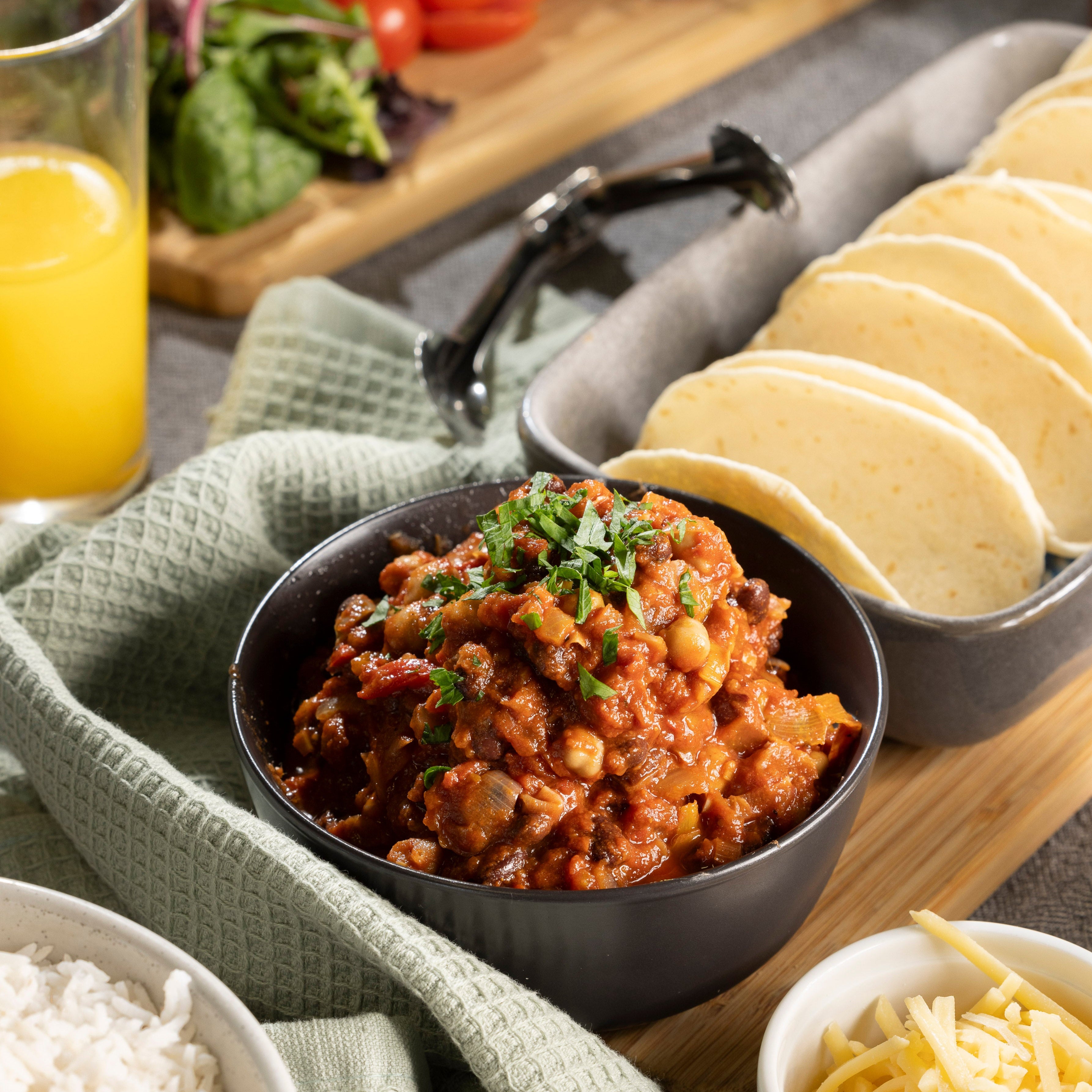 A bowl of vegetarian bean chilli sits on a table with some tortillas and grated cheese.