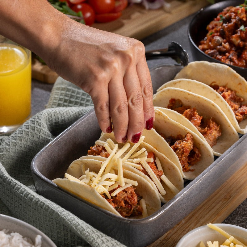 Five soft tacos with slow roast Mexican pork sit in a dish. Someone is sprinkling grated cheese onto the tacos.