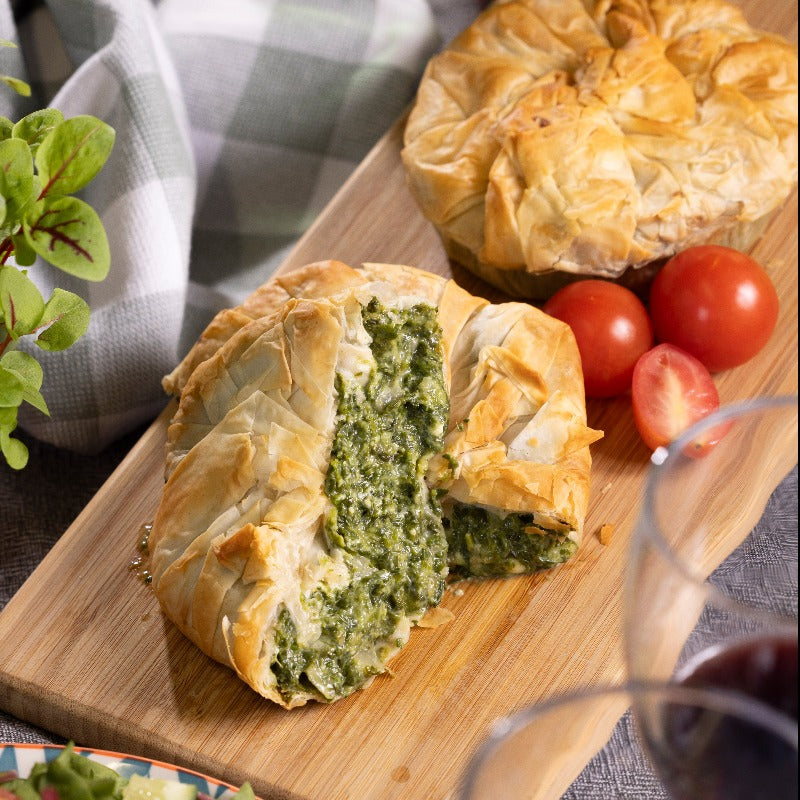 Two spinach & feta pies on a board. There are two wine glasses and a salad in the foreground.