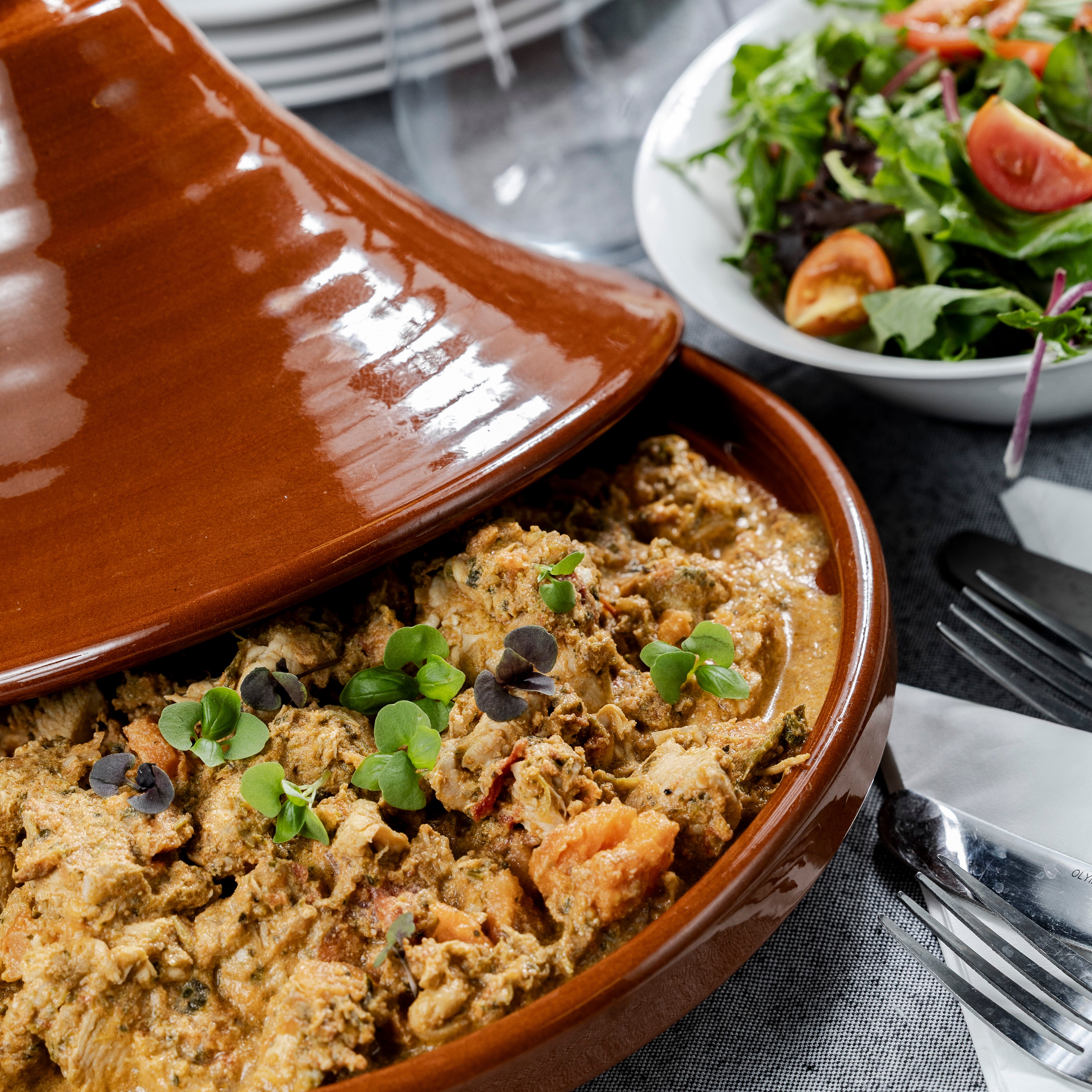 A dish of Moroccan Afro chicken sits in a tagine on a table. There is a bowl of salad in the background.