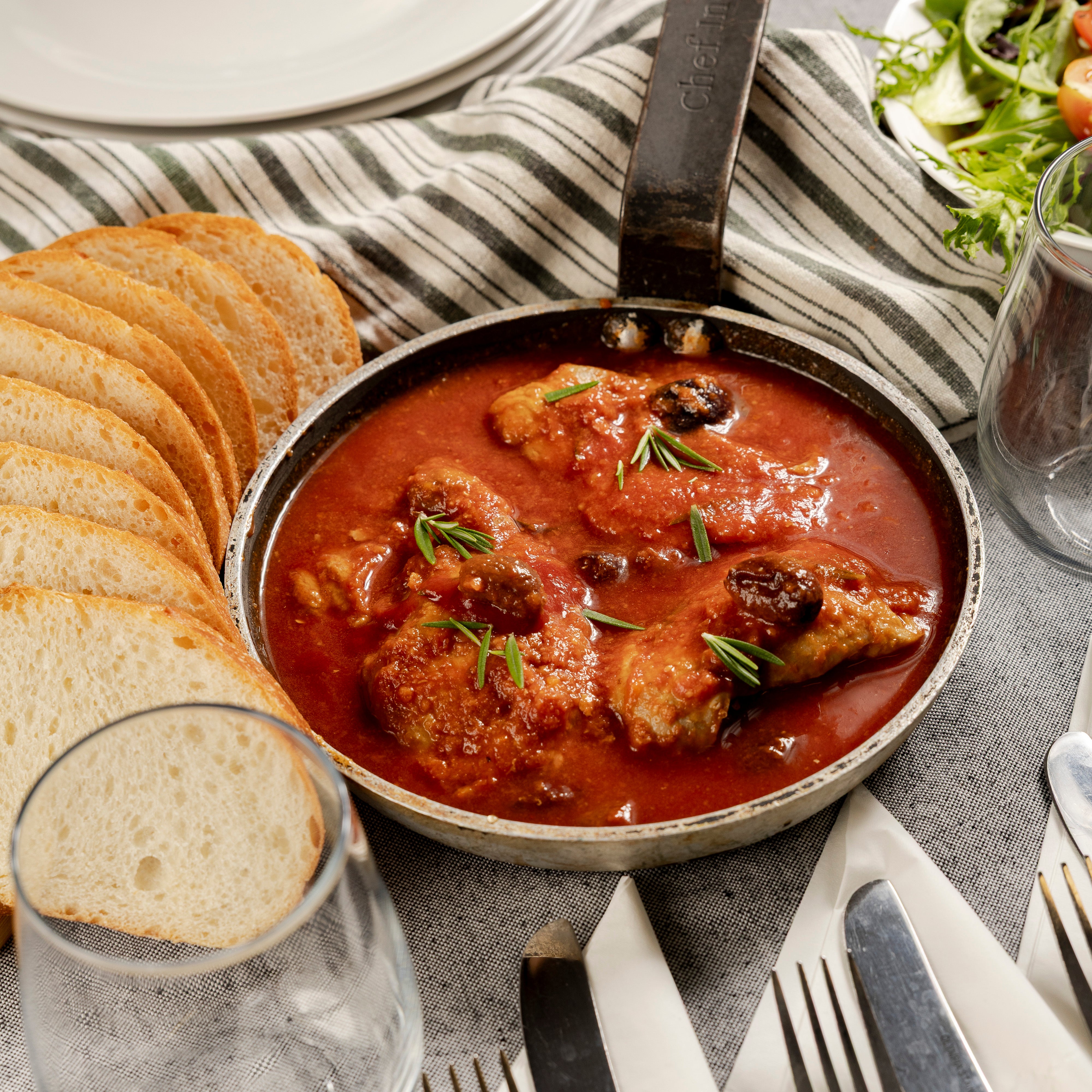 A pot with Chicken Cacciatore on a table. There is a loaf of sourdough bread nearby and a garden salad in the background.