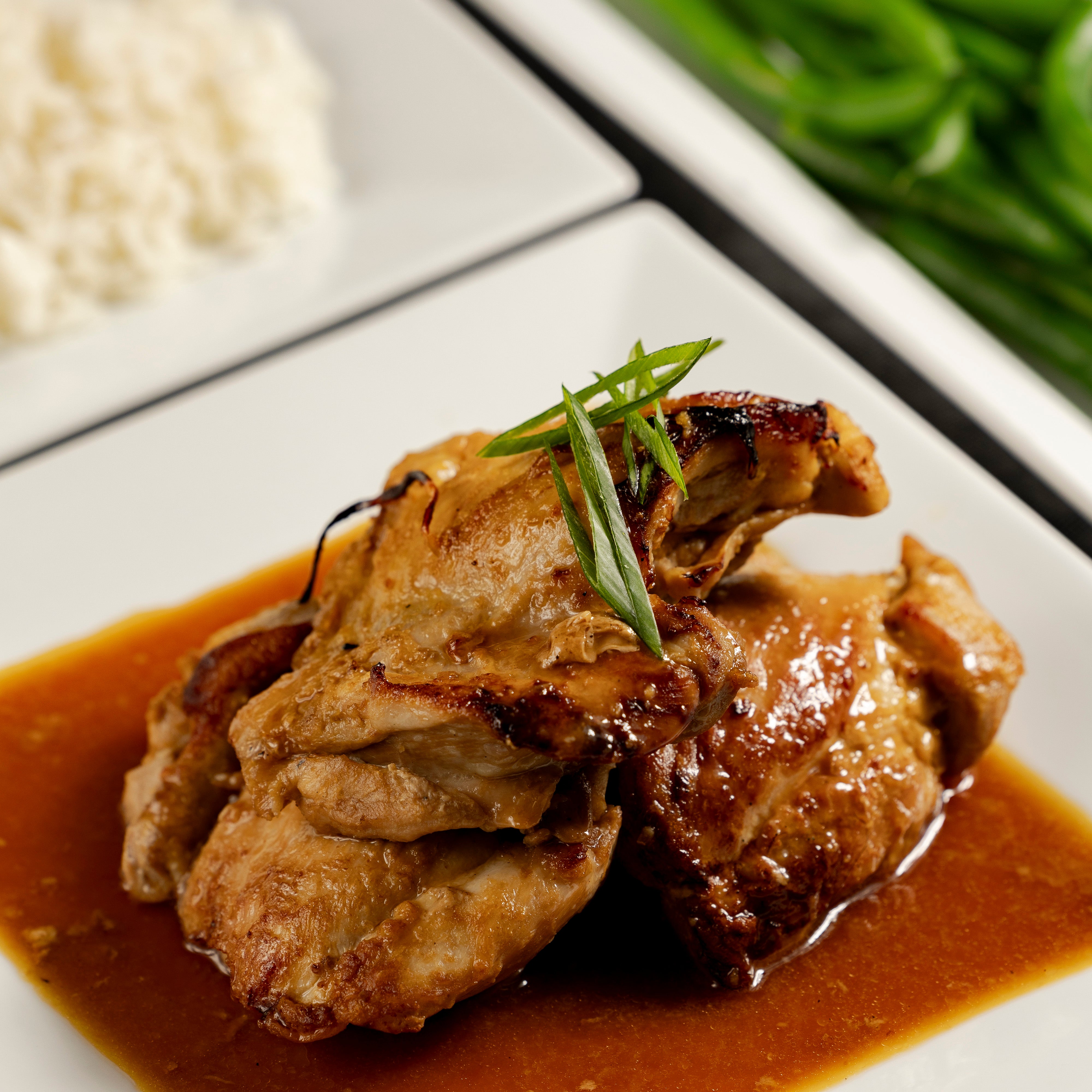 A plate of Teriyaki Chicken with sauce. There is a dish of steamed rice and green beans in the background.