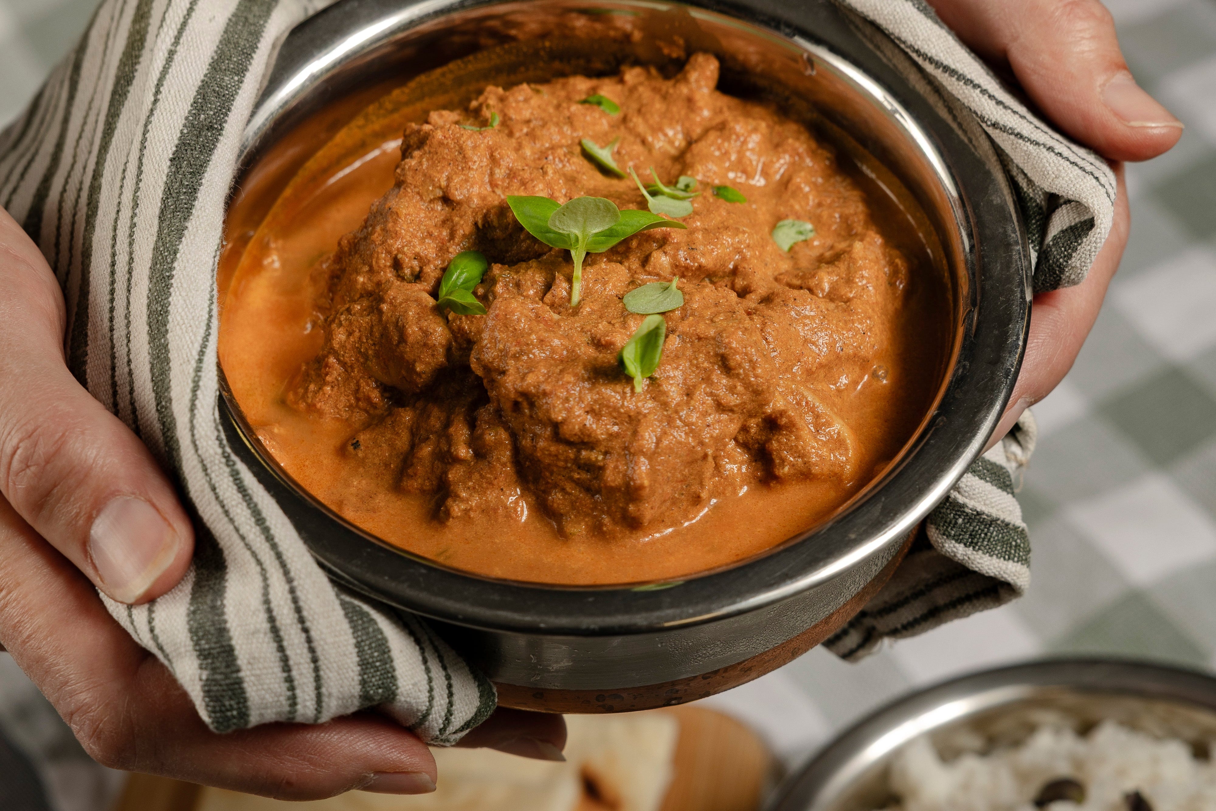Person holding a bowl of Butter Chicken with naan and rice on a checkered tablecloth