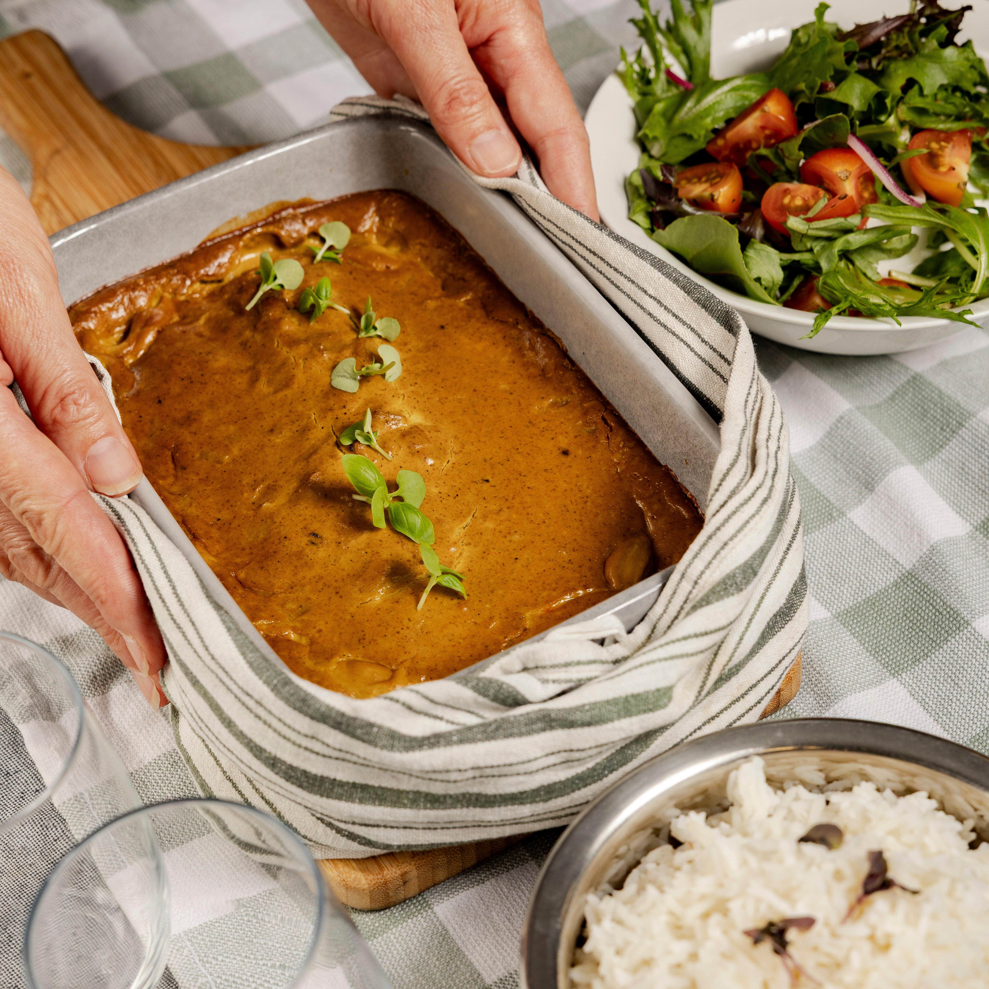 A dish of just baked Sri Lankan fish curry with a green leafy garnish on a checkered tablecloth with a salad and rice bowl.