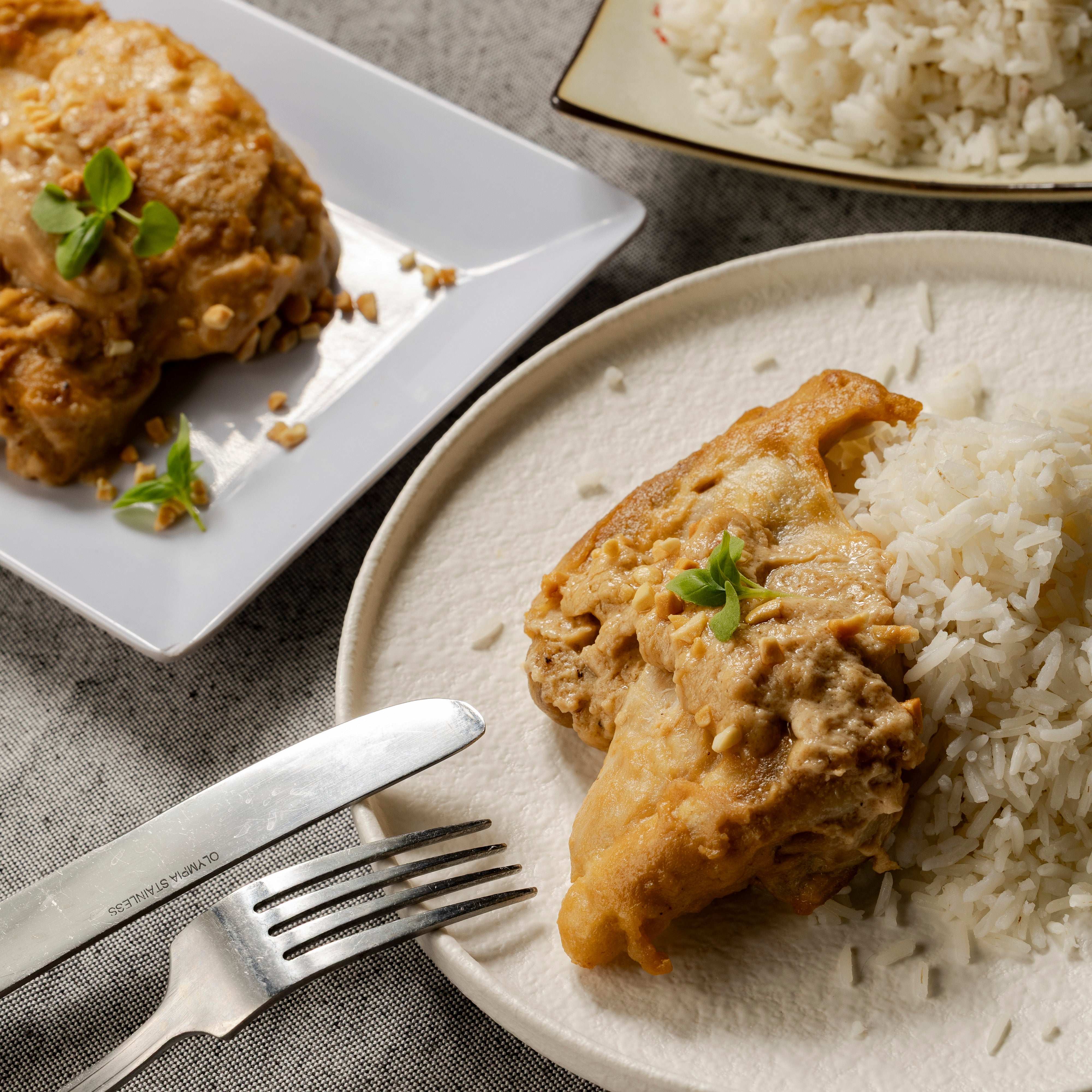 A plate of Satay Chicken and rice sit on a table with a knife & fork leaning up against the plate. There is a platter of Satay Chicken & a dish of rice in the background.