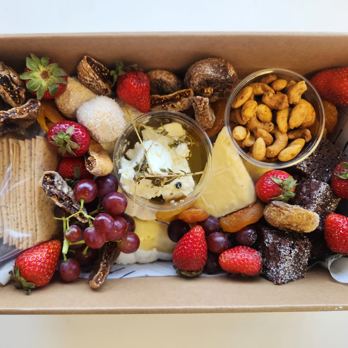 Cheeses & chocolate grazing box with strawberries, grapes, crackers, and spiced nuts in a cardboard box.